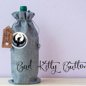 May include: A gray wine bottle bag with a drawstring closure and a round badge featuring a black bird design. A small tag reads "Thank you for visiting my shop". The words "Bad Kitty Buttons" are written in cursive.