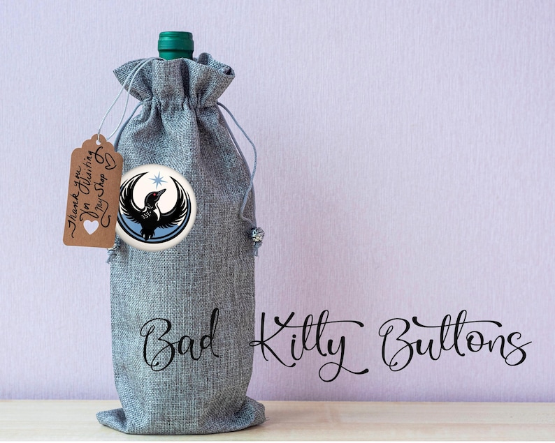 May include: A gray wine bottle bag with a drawstring closure and a round badge featuring a black bird design. A small tag reads "Thank you for visiting my shop". The words "Bad Kitty Buttons" are written in cursive.