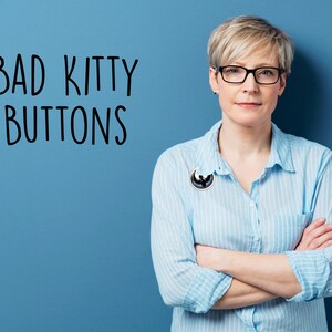 May include: A woman wearing a light blue button-down shirt with a black and white pin. The text "BAD KITTY BUTTONS" is written in black on a blue background. She is wearing black glasses and has her arms crossed.