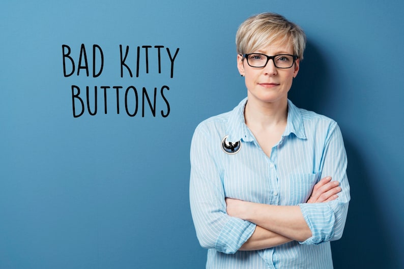 May include: A woman wearing a light blue button-down shirt with a black and white pin. The text "BAD KITTY BUTTONS" is written in black on a blue background. She is wearing black glasses and has her arms crossed.