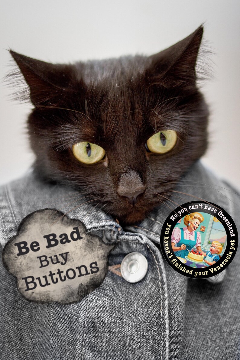 Op de afbeelding: Een zwarte kat met gele ogen draagt een spijkerjasje. De kat heeft twee knopen op de jas en twee ronde spelden. E&eacute;n speld zegt "Be Bad Buy Buttons" en de andere zegt "No you can't have Greenland yet you haven't finished your Venezuela".
