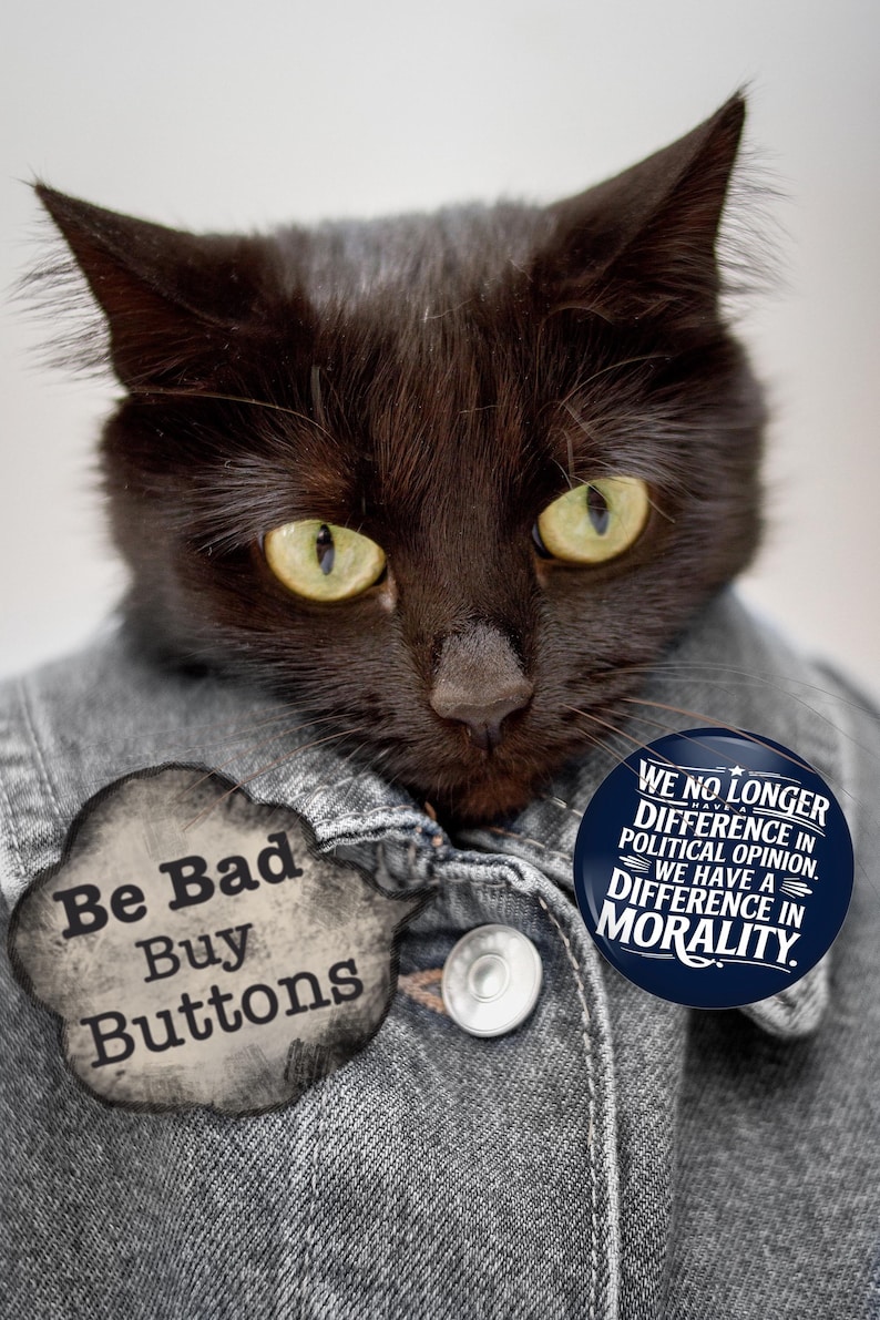 May include: A black cat wearing a denim jacket with two buttons. The cat has yellow eyes and is wearing two buttons. One button says "Be Bad Buy Buttons" and the other says "We no longer difference in political opinion. We have a difference in morality."