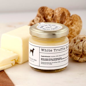 May include: A jar of White Truffle Butter with a gold lid, a block of butter, and several truffles. The jar label includes the text "White Truffle Butter" and "Truffles USA". The butter is a pale yellow colour.