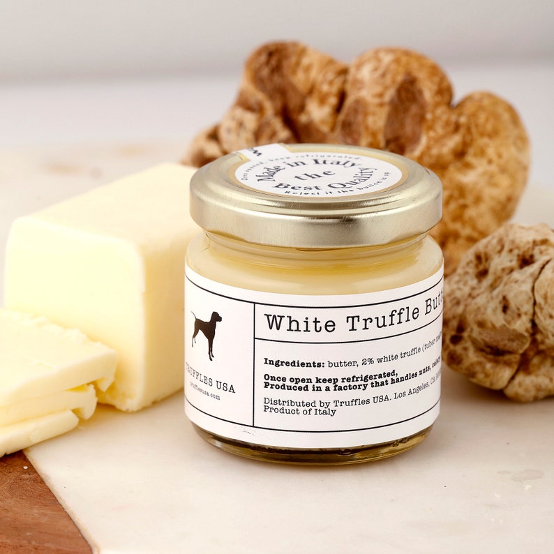 May include: A jar of White Truffle Butter with a gold lid, a block of butter, and several truffles. The jar label includes the text "White Truffle Butter" and "Truffles USA". The butter is a pale yellow colour.