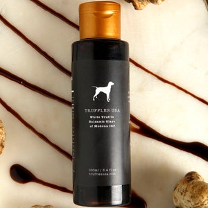 May include: A dark bottle of White Truffle Balsamic Glaze from Truffles USA, with a gold cap, sits on a marble surface. The label features a white dog silhouette. The bottle is surrounded by truffle pieces and balsamic glaze drizzles.