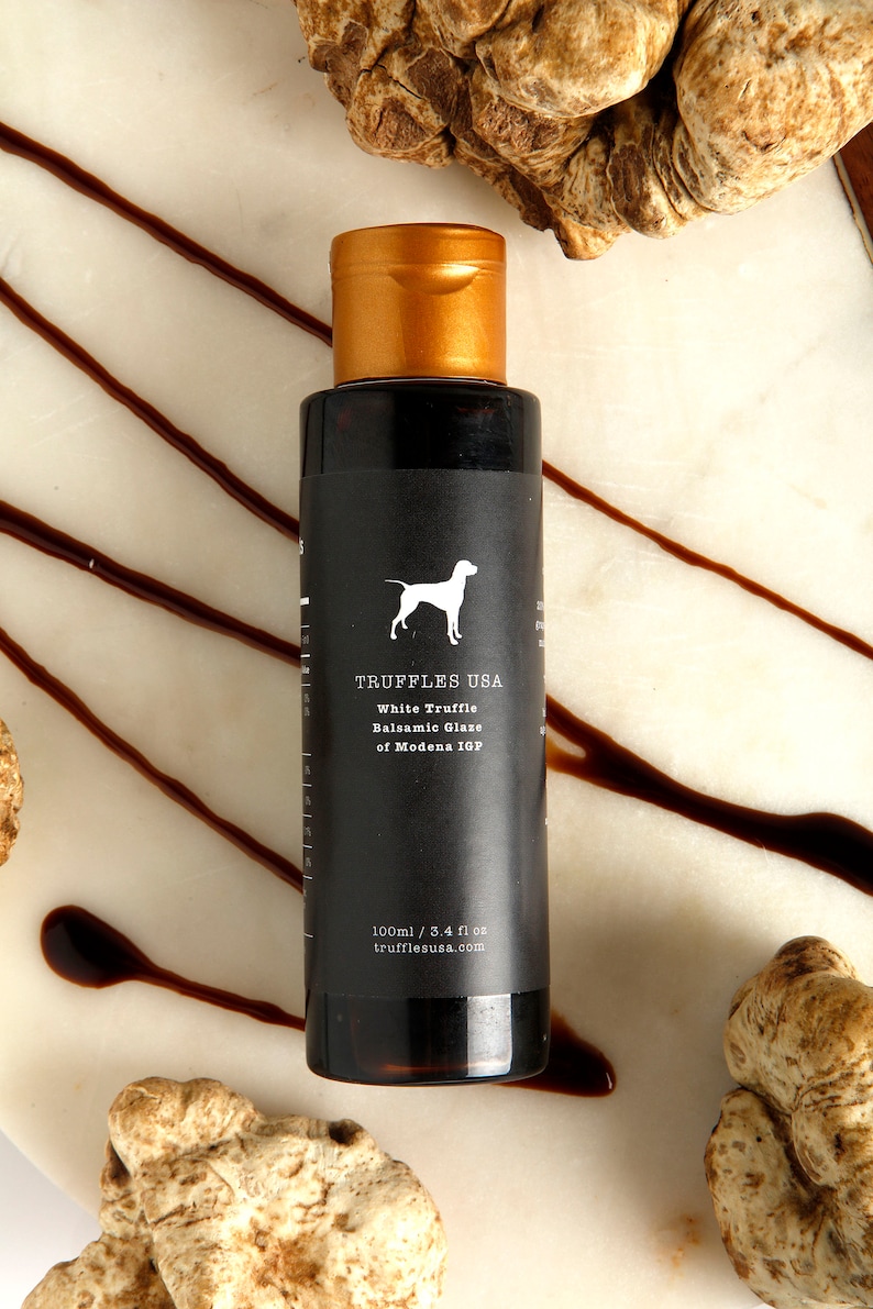 May include: A dark bottle of White Truffle Balsamic Glaze from Truffles USA, with a gold cap, sits on a marble surface. The label features a white dog silhouette. The bottle is surrounded by truffle pieces and balsamic glaze drizzles.