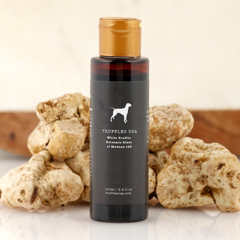 May include: A bottle of White Truffle Balsamic Glaze of Modena IGP from TRUFFLES USA, with a gold cap and a black label featuring a dog silhouette. The bottle is surrounded by several white truffles. The bottle contains 3.4 fl oz.
