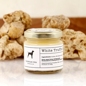May include: A jar of White Truffle Butter with a gold lid, in front of several whole truffles. The label features a dog silhouette and the text "TRUFFLES USA". The ingredients are listed on the label. The product is from Italy.