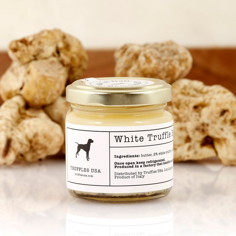 May include: A jar of White Truffle Butter with a gold lid, in front of several whole truffles. The label features a dog silhouette and the text "TRUFFLES USA". The ingredients are listed on the label. The product is from Italy.