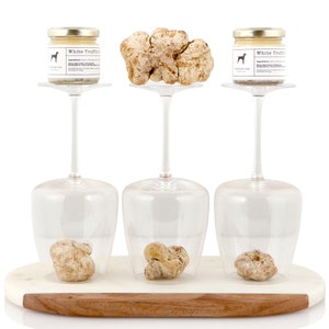 May include: A presentation of white truffles and truffle products. Two small jars of white truffle cream and three whole truffles are displayed on a marble and wood serving board. The jars have gold lids and labels with a dog logo. The truffles are beige.