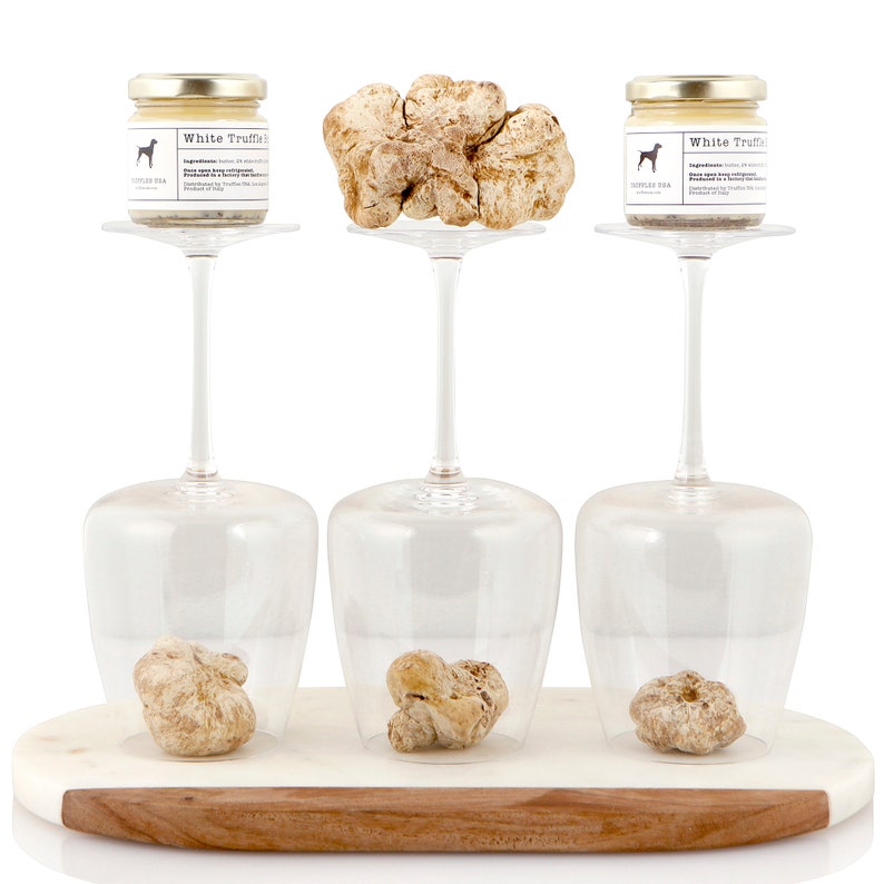 May include: A presentation of white truffles and truffle products. Two small jars of white truffle cream and three whole truffles are displayed on a marble and wood serving board. The jars have gold lids and labels with a dog logo. The truffles are beige.