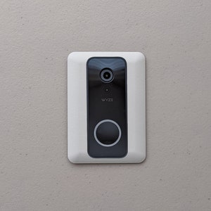 May include: A white rectangular smart doorbell with a black center panel. The black panel has a camera lens, the word "WYZE", and a large circular button. The doorbell is mounted on a light gray wall.