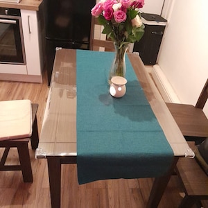May include: A teal table runner on a wooden table with a clear glass top. A vase of pink and white roses sits on the table with a white ceramic candle holder.