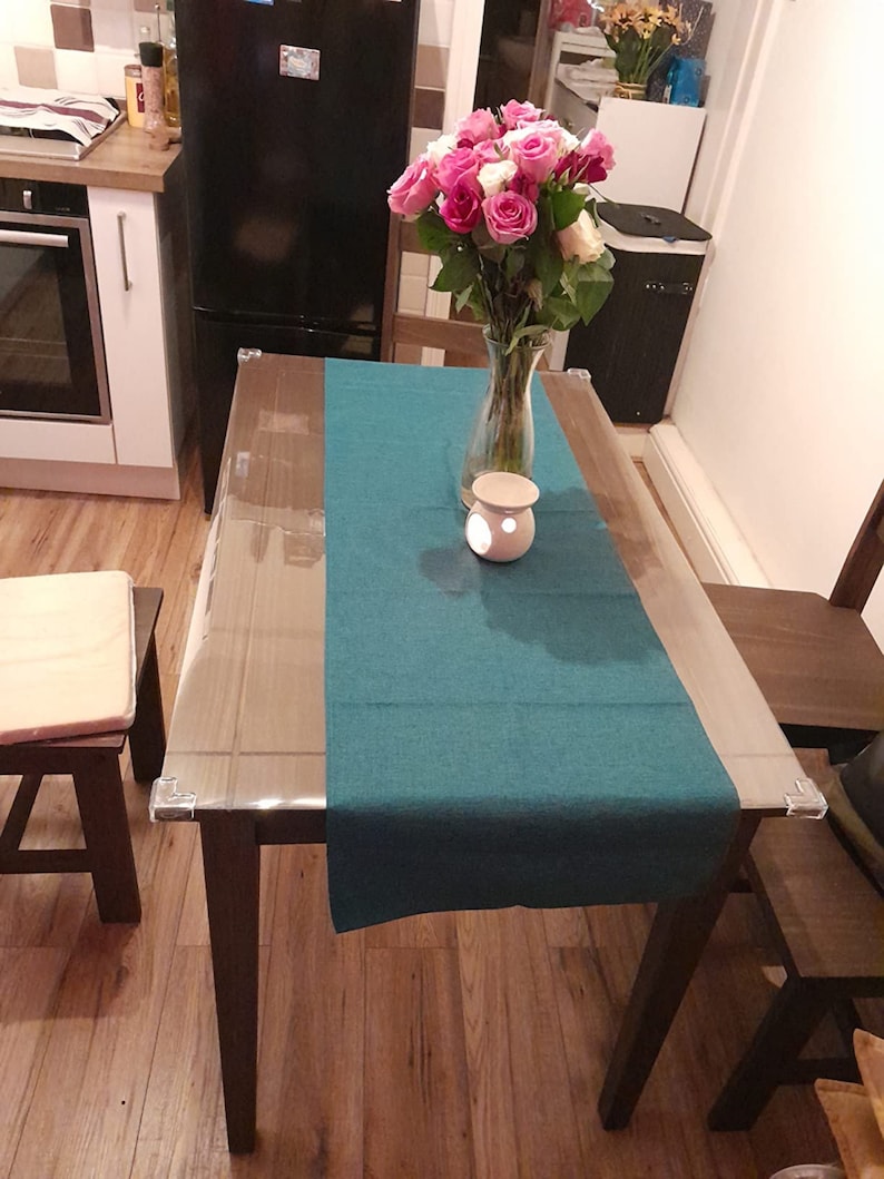 May include: A teal table runner on a wooden table with a clear glass top. A vase of pink and white roses sits on the table with a white ceramic candle holder.