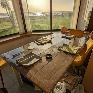 May include: A wooden table with a clear glass top, set with four placemats and coasters. The table is positioned in front of a large window with a view of the ocean and a grassy area.