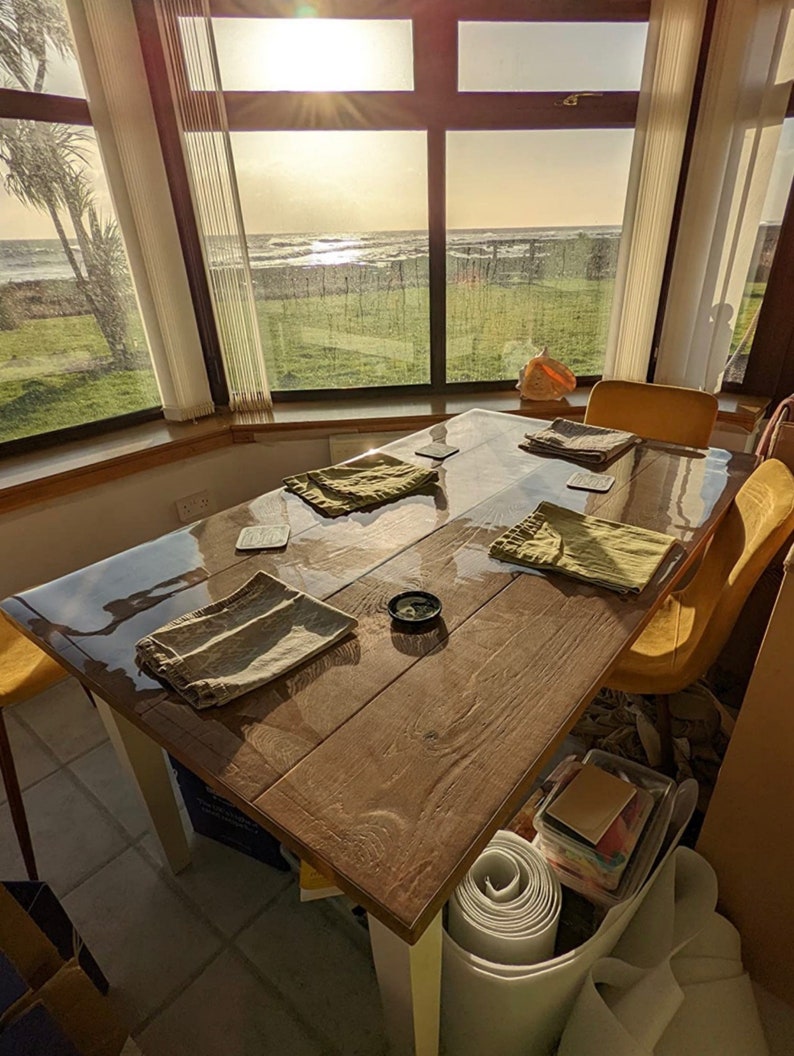 May include: A wooden table with a clear glass top, set with four placemats and coasters. The table is positioned in front of a large window with a view of the ocean and a grassy area.