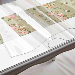 May include: A clear plastic table protector is rolled up on a table with a floral tablecloth. The protector is designed to protect the table surface from scratches and spills.