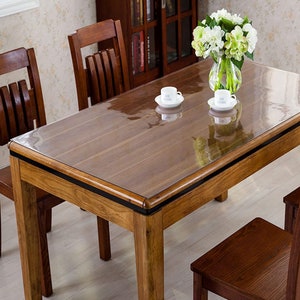 May include: A clear plastic table protector placed on top of a wooden table. The table protector is designed to protect the table surface from scratches and spills. The table is brown and has a black border. There are two white cups and saucers on the table, and a vase with white flowers.