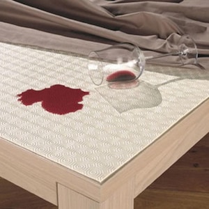 May include: A white absorbent paper towel sheet with a repeating geometric pattern lies flat on a light wood surface. A clear glass wine glass is tipped over on the paper, with a large, irregular red wine stain visible on the paper surface.