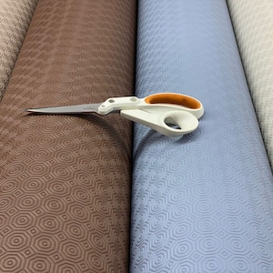 May include: Rolled-up, textured vinyl tablecloths in shades of beige, brown, and blue. A pair of white and orange handled scissors rests on the brown roll. The tablecloths have a repeating geometric pattern.