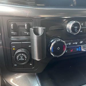 May include: A black plastic phone holder mounted in a car's dashboard. The holder has a textured surface and a circular opening at the top. The holder is attached to the dashboard with a screw.