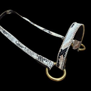 Puede incluir: Cabezada decorativa con estampado de piel de serpiente en negro, blanco y marrón. La cabezada tiene anillos de metal dorados y está adornada con pequeños diamantes de imitación transparentes. El fondo es negro sólido.