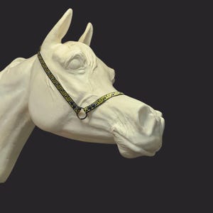 Puede incluir: Una escultura de cabeza de caballo blanca con un cabestro decorativo negro y dorado. El cabestro presenta un patrón ornamental y repetitivo. La escultura está sobre un fondo oscuro.