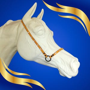 Puede incluir: Una estatua de cabeza de caballo blanca con un cabestro decorativo naranja y blanco. El cabestro tiene detalles en negro. El fondo es un degradado azul con elementos decorativos dorados.