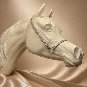 Puede incluir: Una escultura de cabeza de caballo color crema con un cabestro decorativo. El cabestro es de color beige con un patrón repetido y detalles en gris oscuro. Un anillo dorado conecta las correas del cabestro.