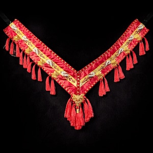 Puede incluir: Collar decorativo rojo con detalles dorados y blancos y borlas rojas. El collar en forma de V presenta un colgante central y probablemente se utiliza con fines ceremoniales o decorativos.
