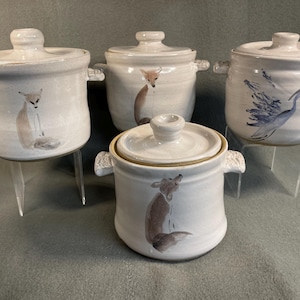 May include: Four white ceramic lidded pots with handles. Each pot features a hand-painted animal design: foxes and a blue heron. The pots are displayed on clear stands, showcasing their unique artwork and craftsmanship.