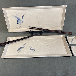May include: A Japanese-style sushi set featuring two rectangular serving platters, two small square sauce dishes, and dark brown chopsticks. The off-white ceramic pieces are decorated with blue crane illustrations.