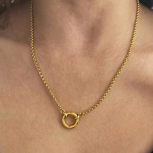 May include: A gold-colored necklace featuring a delicate chain and a circular pendant. The necklace is worn around the neck, with the pendant resting near the collarbone. The chain has a woven design, and the pendant is a simple, polished circle.