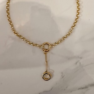 Puede incluir: Un collar dorado con un delicado diseño de cadena. El collar presenta una cadena circular que forma un bucle, con un pequeño colgante colgando del centro. El colgante tiene un pequeño anillo en la parte superior y otro más pequeño en la parte inferior.