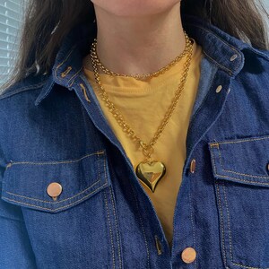 May include: A gold chain necklace with a large, heart-shaped pendant. The necklace is layered over a yellow t-shirt and a denim jacket.
