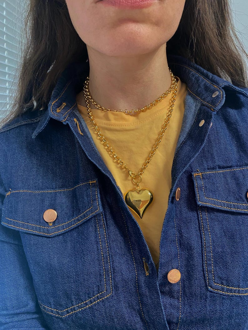 May include: A gold chain necklace with a large, heart-shaped pendant. The necklace is layered over a yellow t-shirt and a denim jacket.