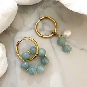 May include: A pair of gold hoop earrings with a charm dangling from one. The charm is a single white pearl with a cluster of light blue beads below it. The other earring has a circle of light blue beads around the hoop.
