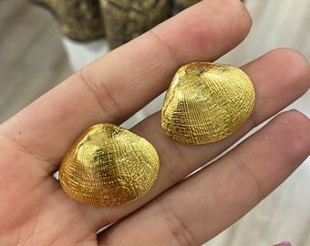 Könnte beinhalten: Zwei goldfarbene Ohrringe in Muschelform. Die Ohrringe haben eine strukturierte Oberfläche, die an die natürlichen Rippen einer Muschel erinnert. Die Ohrringe sind rund und haben ein glänzendes, metallisches Finish. Die Ohrringe sind ein Schmuckstück.