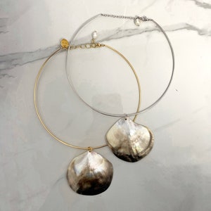 Puede incluir: Dos collares con colgantes de concha. Uno es dorado, el otro plateado. Ambos tienen un diseño circular con un gran colgante de concha iridiscente. Los collares se exhiben sobre una superficie de mármol.