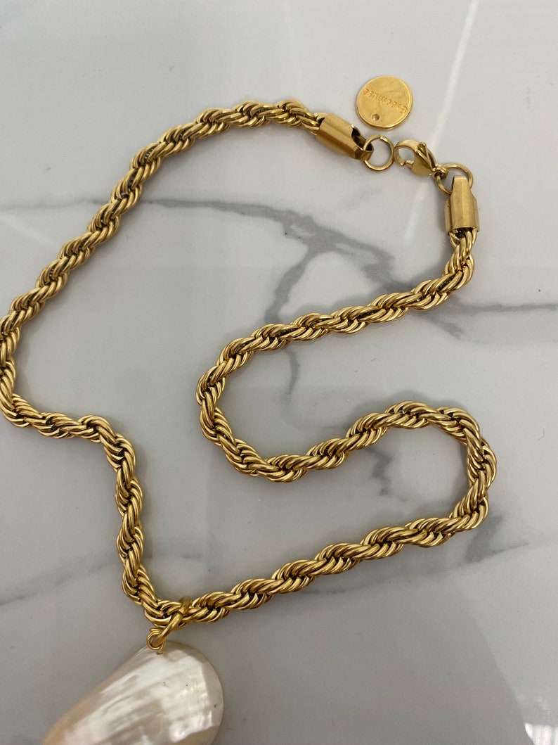 May include: A gold rope chain necklace with a large white pearl pendant. The chain has a lobster clasp and a small round tag with the word "remarkable" on it.