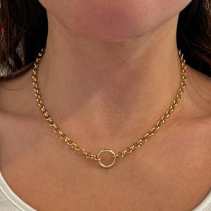 May include: Gold-toned necklace featuring a chain with a circular pendant. The necklace is made of a shiny, yellow metal and is worn around the neck. The chain links are a medium size and the pendant is a simple, smooth circle.