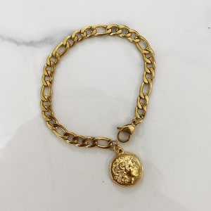 May include: A gold chain bracelet with a gold coin charm featuring a profile of a person's face.