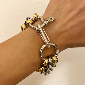 Könnte beinhalten: Ein silbernes und goldenes Kettenarmband mit einem großen silbernen Verschluss. Das Armband besteht aus mehreren Kettengliedern in verschiedenen Größen und Stilen.