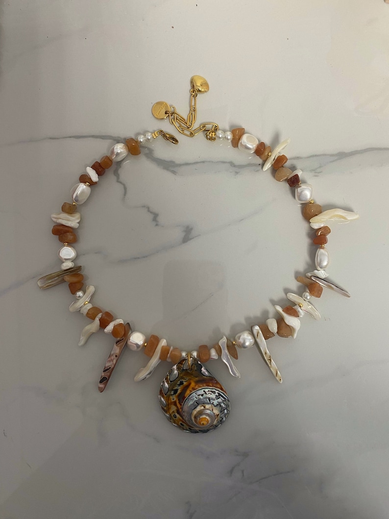 May include: A gold chain necklace with a large seashell pendant. The necklace is made with brown and white stones and has a gold clasp.