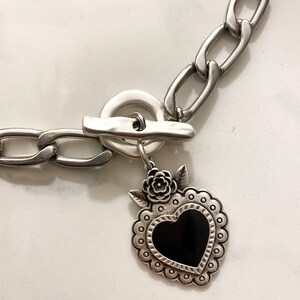 May include: A silver chain necklace with a silver toggle clasp and a silver heart pendant with a black enamel center. The heart pendant has a floral design around the edge.