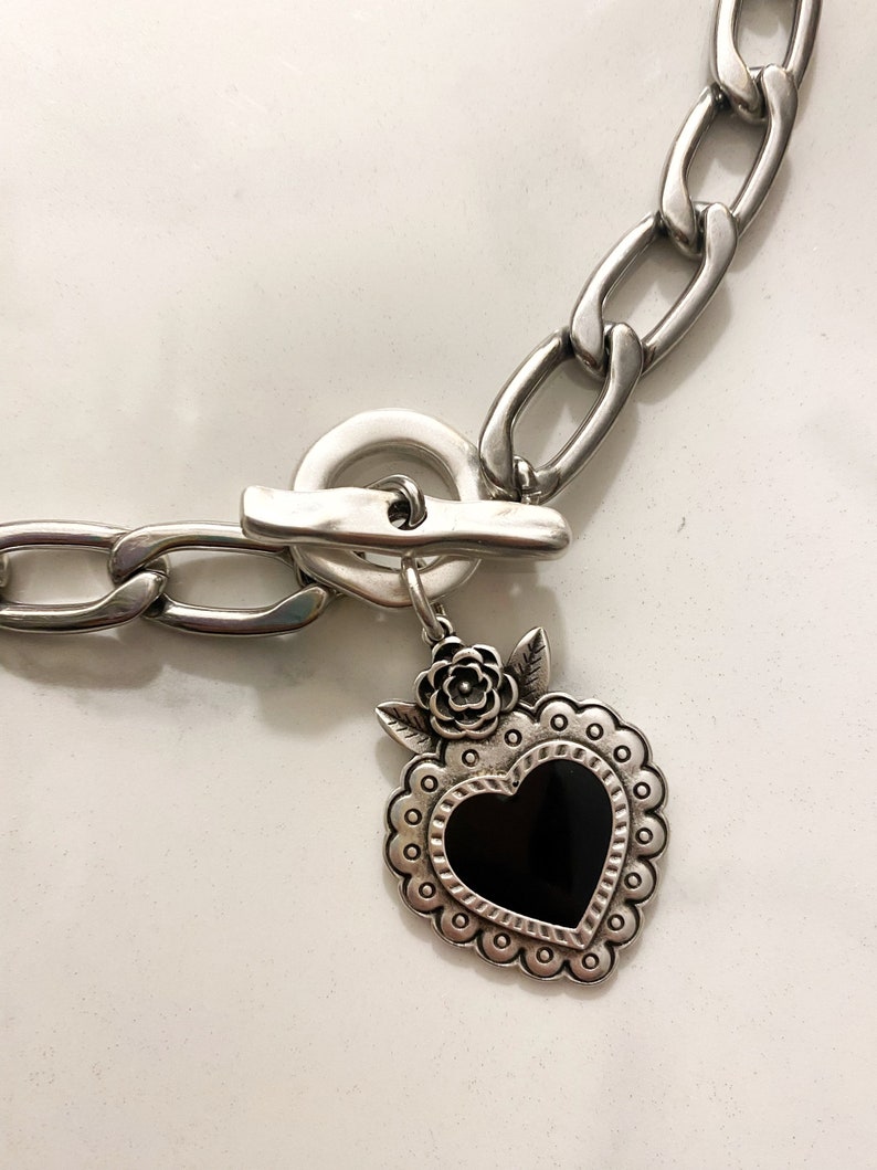 May include: A silver chain necklace with a silver toggle clasp and a silver heart pendant with a black enamel center. The heart pendant has a floral design around the edge.