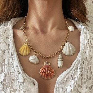 Op de afbeelding: Een gouden ketting met meerdere schelpbedels. De bedels zijn in verschillende kleuren, waaronder wit, geel en oranje. De ketting wordt gedragen door een persoon met een wit kanten topje.