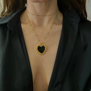 Pu&ograve; includere: Una collana d'oro con un ciondolo a forma di cuore nero. Il ciondolo ha un design delicato e decorato.
