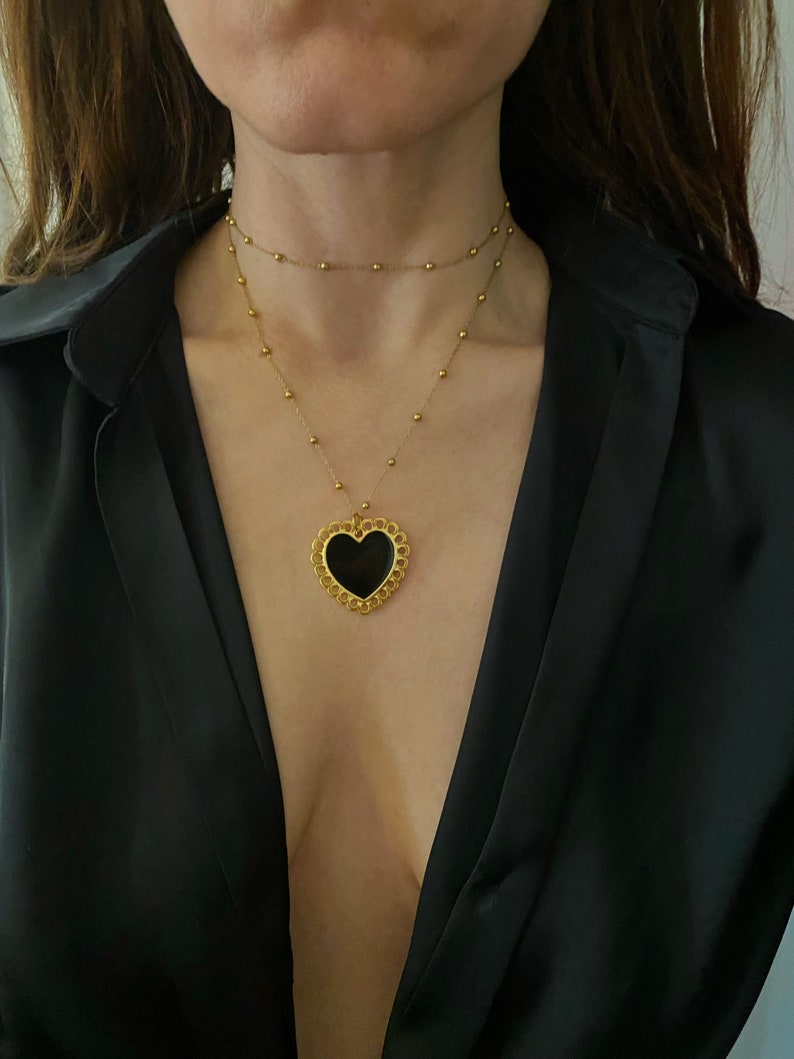Pu&ograve; includere: Una collana d'oro con un ciondolo a forma di cuore nero. Il ciondolo ha un design delicato e decorato.