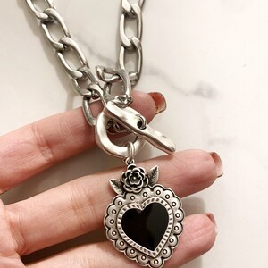 May include: Silver chain necklace with a large silver heart pendant. The heart has a black center and is decorated with a silver flower and leaves.